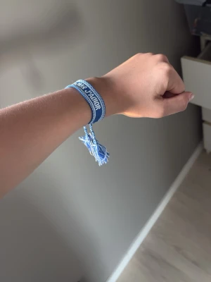 Blått Christian Dior J'Adior armband - Snyggt blått armband från Christian Dior med texten 'J'Adior' broderad i vitt. Armbandet är gjort i tyg och har justerbara tofsar för perfekt passform. Perfekt accessoar för att lyfta din outfit med en lyxig touch.