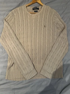 Beige tröja från Polo Ralph Lauren - Äkta kabelstickad tröja i beige från Polo Ralph Lauren med rund halsringning och diskret broderad logga på bröstet. Tröjan har långa ärmar och ribbade muddar vid ärmslut och nederkant. Perfekt för lager-på-lager och en klassisk look. Priset kan diskuteras, storlek M hör gärna av er efter frågor.