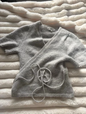 Grå omlottstickad tröja - Mysig grå stickad omlott-tröja med korta ärmar och knytband i midjan. Tröjan har en klassisk v-ringning och är tillverkad i ett mjukt, fluffigt material som känns skönt mot huden. Perfekt för lager-på-lager och enkel att styla till olika outfits.