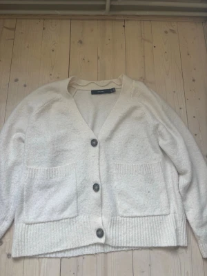 Vit kofta från Vero Moda - Mysig vit kofta från Vero Moda med stora knappar framtill och två rejäla fickor. Koftan har en avslappnad passform, ribbade muddar och är perfekt att slänga på sig när det är kallt ute. Materialet är mjukt och fluffigt för extra komfort.