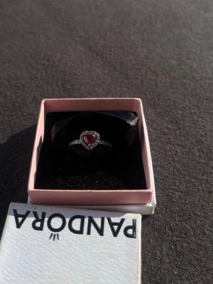 Pandora silverring med hjärtformad sten - Snygg silverring från Pandora med en röd hjärtformad sten i mitten, omgiven av små gnistrande stenar. Ringen har en klassisk och romantisk design som verkligen sticker ut. Perfekt accessoar för att lyfta din stil. Det finns tecken på användning men inget som syns när den används. Storlek 16