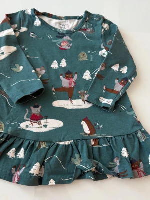 Grön  klänning med vintermotiv  - Supersöt grön  klänning  med vintermönster av djur som åker skridskor och skidor. Klänningen har volangkant nedtill, långa ärmar och knappar vid axeln. Perfekt för mysiga vinterdagar.