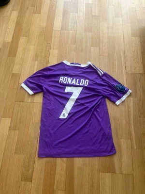 Real Madrid Ronaldo 7 lila fotbollströja (S) - Säljer en lila Real Madrid fotbollströja med Ronaldo och nummer 7 på ryggen. Tröjan har vita detaljer, korta ärmar och officiella klubbmärken samt sponsortryck på framsidan. Perfekt för fans av Ronaldo och Real Madrid.