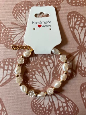 Pärlarmband med blommor och gulddetaljer - Snyggt armband med vita pärlor och små blommor i plast, varvat med guldfärgade pärlor. Armbandet har en justerbar kedja och ger en söt och trendig look. Perfekt accessoar för att lyfta din outfit.