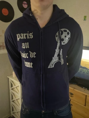 Mörkblå hoodie blinglucid - Mörkblå hoodie med dragkedja och huva. Framsidan har ett glittrigt tryck med Eiffeltornet, måne och texten 'paris au clair de lune' i silver. Mjuk bomullskänsla och känguruficka framtill. Perfekt för dig som gillar unika detaljer och Paris-vibes.