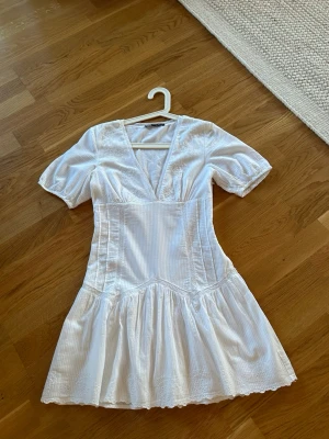  Vit kortklänning med v-ringning - Superfin vit kortklänning med v-ringning och korta puffärmar. Klänningen har markerad midja, volangig kjol och diskreta broderade detaljer. Perfekt för sommarens alla tillfällen och ger en fräsch och romantisk look.