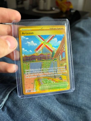 Pokémonkort Artazon Stadium Gold - Säljer ett Pokémonkort av typen Stadium Trainer med namnet Artazon. Kortet är en specialutgåva med guldfärgad ram och holografisk yta, motivet visar en färgglad väderkvarn och byggnad. Kortet ligger i ett skyddande plastfodral.