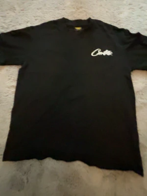 Svart Corteiz t-shirt med tryck - Svart t-shirt från Corteiz med vit logga tryckt på bröstet. Klassisk passform och rund halsringning, perfekt för en clean streetwear-look. Materialet känns mjukt och skönt mot huden.