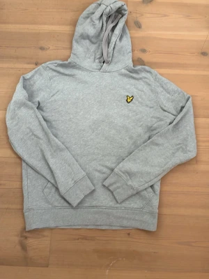 Grå hoodie från Lyle & Scott - Snygg grå hoodie från Lyle & Scott med klassisk gul logga på bröstet. Hoodien har huva med dragsko, ribbade muddar och magficka. Perfekt för en avslappnad och stilren look.