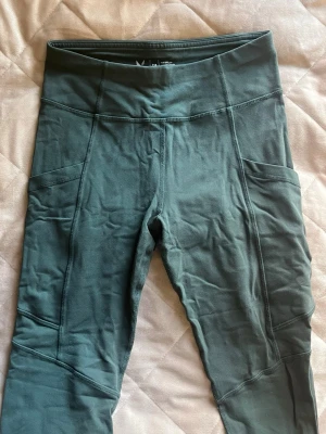 Gröna träningstights från SOC - Snygga gröna träningstights från SOC med hög midja och praktiska sidofickor. Tillverkade i ett stretchigt material som andas, perfekt för gymmet eller löprundan. Tightsen har synliga sömmar och sitter skönt på kroppen.