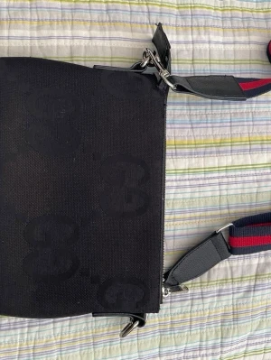 Svart axelväska från Gucci - Snygg svart axelväska från Gucci med GG-monogram i tyg. Väskan har en bred axelrem i rött och blått med läderdetaljer och silverfärgade metalldelar. Perfekt för dig som vill ha en stilren och trendig accessoar.