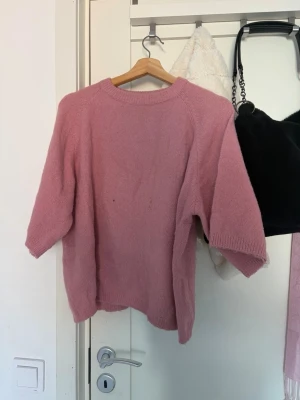 Rosa stickad tröja från 157 - Mysig rosa stickad tröja från 157 med rund halsringning och halvlånga ärmar. Tröjan har en enkel och avslappnad passform, perfekt att slänga på sig när du vill vara både bekväm och snygg. Ribbstickade detaljer vid hals, ärmslut och nederkant.