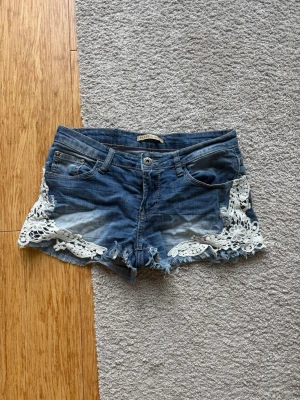 Blå jeansshorts med spetsdetaljer - Snygga blå jeansshorts från Rue21 med slitna kanter och vita spetsdetaljer på sidorna. Klassisk femficksmodell med knapp och dragkedja framtill. Perfekta för en chill sommarlook.