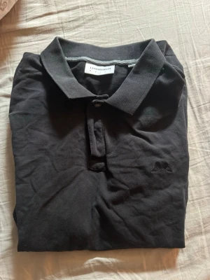 Svart pikétröja från Lindbergh - Klassisk svart pikétröja från Lindbergh med krage och knappslå framtill. Tröjan har korta ärmar och är tillverkad i mjuk bomull. Perfekt för en stilren och avslappnad look.