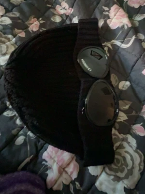 Svart mössa från C.P. Company - Säljer en svart ribbstickad mössa från C.P. Company med de ikoniska inbyggda goggles framtill. Mössan är i ett mjukt och stretchigt material som sitter skönt på huvudet. Perfekt för dig som vill sticka ut med en streetwear-inspirerad look. Köpare står för frakt