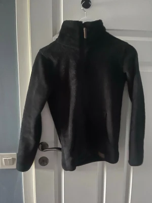 Svart fleece tröja med halv dragkedja - Mysig svart fleece tröja med hög krage och halv dragkedja framtill. Perfekt för kalla dagar och enkel att dra på över en t-shirt. Tröjan har långa ärmar och en mjuk, fluffig känsla. Passar dig som gillar enkel och stilren design.