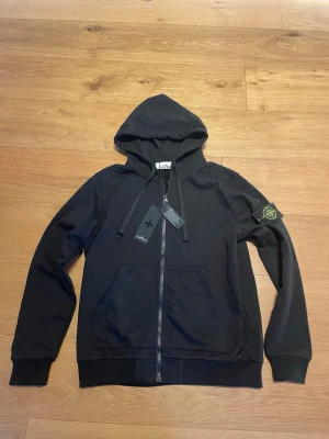 Svart hoodie från Stone Island - Svart hoodie från Stone Island med dragkedja framtill, två fickor och klassisk logopatch på vänster ärm. Tillverkad i mjukt bomullsmaterial och har justerbar huva med snören. Perfekt för en avslappnad och stilren look.