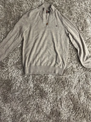 Grå merinoulltröja från Morris - Snygg beige tröja från Morris i 100% merinoull med halv dragkedja och liten broderad logga på bröstet. Tröjan har ribbade muddar och hög krage, perfekt för en stilren look.