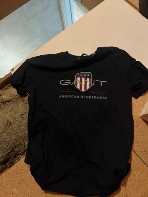 Svart GANT t-shirt med tryck - Svart t-shirt från GANT med klassiskt tryck framtill som visar logga och texten 'American Sportswear'. T-shirten har rund hals och korta ärmar. Perfekt för en avslappnad och sportig stil.