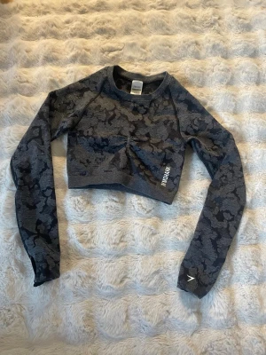 Mörkgrå crop top från Gymshark - Snygg mörkgrå långärmad crop top från Gymshark med camomönster. Tröjan har tight passform, tumhål vid ärmsluten och diskret logga på bröstet och ärmen. Perfekt för gymmet eller träning. Materialet är stretchigt och andas bra.
