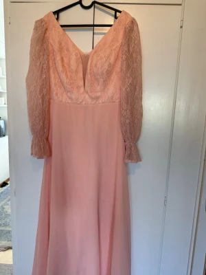 Lång rosa klänning med spetsärmar - Säljer en elegant långklänning i ljusrosa med v-ringning och långa spetsärmar. Klänningen har en flowy kjol och romantiska detaljer vid ärmsluten. Perfekt för dig som vill sticka ut med en feminin och klassisk look.