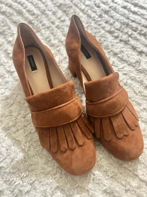 Bruna mockaloafers från Mango - Snygga bruna loafers i mocka från Mango med dekorativa fransar framtill och låg klack. Skorna har en klassisk form och är perfekta för dig som gillar en stilren och trendig look.