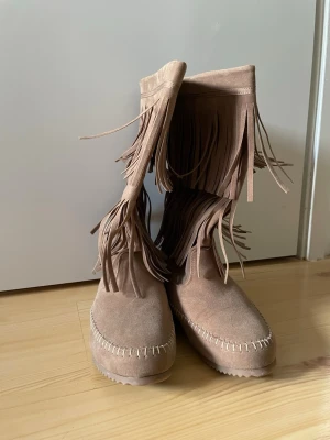 Beiga mockastövlar med fransar - Säljer ett par höga stövlar i storlek 38 i beige mocka med dubbla rader fransar och dekorativa sömmar framtill. Stövlarna har en mjuk och bekväm känsla och ger en boho-vibe till outfiten. 