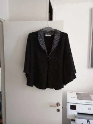 Svart ponchokappa med grå krage - Ponchokappa i svart med bred gråmelerad krage och knäppning framtill. Kappan har en lös och fladdrig passform med vida ärmar, perfekt för lager-på-lager. Materialet ser ut att vara mjukt och stickat, vilket ger en mysig känsla.