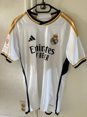 Real Madrid matchtröja Vini Jr. 7 - Snygg vit Real Madrid matchtröja från Adidas med Vini Jr. och nummer 7 på ryggen. Tröjan har svarta och guldiga detaljer, klubbmärke på bröstet och sponsortryck framtill. Korta ärmar och klassisk sportig passform, perfekt för fotbollsfans.