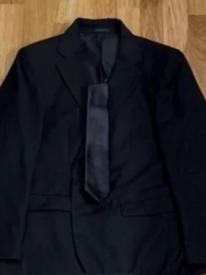 Svart kavaj från Dressmann - Storlek 52 - Klassisk svart kavaj som passar lika bra till kontoret som till mer formella tillfällen! Storlek: 52. Regular fit. Varumärke: Dressmann. Klassisk krage och knäppning framtill. Tre praktiska innerfickor samt tre fickor på framsidan. Material: Yttertyg: 80% Polyester och 20% Viskos. Foder: 100% Polyester. Ärmar: 100% Polyester. Säljs i mycket bra skick! Slips på bild ingår ej. Kika gärna in på mina andra annonser, jag säljer flera klädesplagg - såsom bland annat ett par matchande kostymbyxor!
