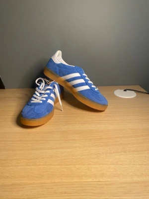 Blå Adidas gazelle  - Snygga blå sneakers från Adidas med klassiska vita ränder på sidorna och vit häl. Ovandelen är i mocka och sulan är i gummi med en brun ton. Skorna har vita skosnören och en retroinspirerad design som sticker ut.