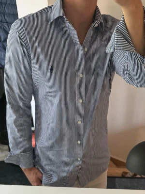 Randig skjorta från Polo Ralph Lauren - Klassisk blåvit randig skjorta från Polo Ralph Lauren med lång ärm och knappar framtill. Skjortan har en broderad logga på bröstet och är tillverkad i bomull. Perfekt för en stilren och avslappnad look. Storlek 39 vilket motsvarar m 