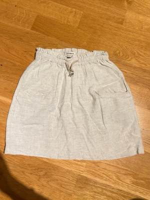 Beiga linnekjol från Pull&Bear - Säljer en ljusbeiga linnekjol från Pull&Bear med avslappnad passform och resår i midjan. Kjolen har snörning i midjan för justerbar passform och är perfekta för varma dagar. Materialet är svalt och luftigt linne.