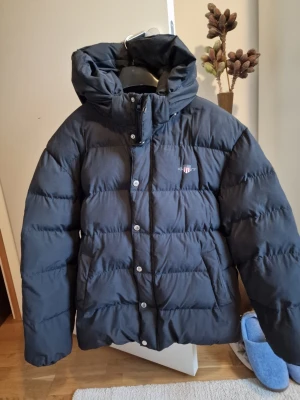 Svart dunjacka från Gant med huva - Snygg svart dunjacka från Gant med stor vadderad huva och knäppning med både dragkedja och tryckknappar. Jackan har en klassisk puffer-modell och Gant-logga broderad på bröstet. Perfekt för kalla vinterdagar.
