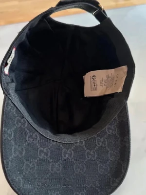 Svart keps från Gucci med GG-mönster - Snygg svart keps från Gucci med klassiskt GG-monogram över hela tyget. Justerbar rem baktill med metallspänne och böjd skärm. Tillverkad i bomullsmix med exklusiv känsla och diskret logga. Perfekt för dig som gillar streetstyle och lyxiga accessoarer.