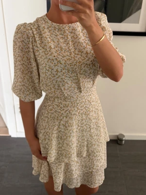 Blommig kortklänning med puffärm - ADOORE strl 38/M - Supersöt kortklänning i ljusbeige med små gula blommor. Klänningen har puffiga korta ärmar, rund halsringning och volangdetalj i kjolen. Perfekt för sommarens alla tillfällen och ger en romantisk vibe. Klänningen funkar till dig i storlek S/M