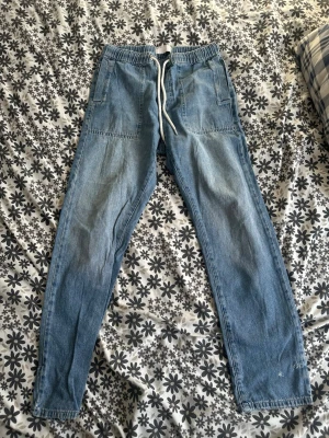 Ljusblå jeans med dragsko från ASOS - Säljer ett par ljusblå jeans från ASOS DESIGN med elastisk midja och vit dragsko. Jeansen har raka ben, stora framfickor och är tillverkade i denim. Perfekta för en avslappnad och trendig stil.