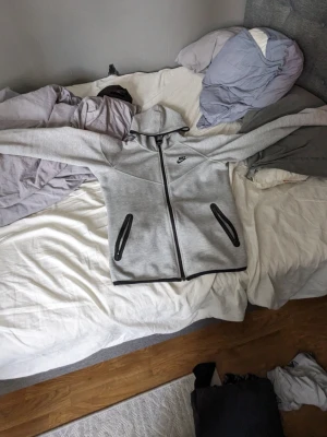 Grå Nike hoodie med dragkedja - Snygg grå hoodie från Nike med svart logga på bröstet och hel dragkedja framtill. Två svarta dragkedjefickor och svart kant nertill. Klassisk huva och sportig look, perfekt till streetwear eller träning.