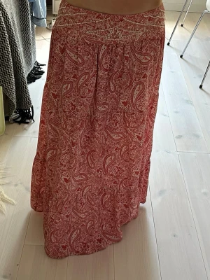 Lågmidjad paisleymönstrad långkjol - Snygg röd långkjol med vitt paisleymönster och dekorativa broderier i midjan. Kjolen har en avslappnad passform och är perfekt för dig som gillar bohemisk stil. Tillverkad i ett mjukt och lätt tyg som känns skönt mot huden.