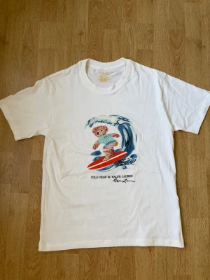 Vit t-shirt Polo Bear Ralph Lauren - Vit t-shirt från Polo Ralph Lauren med en färgglad print av en surfande björn på bröstet. Modellen är custom slim fit och tillverkad i skön bomull. Perfekt för en avslappnad och cool stil med en lekfull touch till sommaren. Den är helt ny och aldrig använd. Skrivs vid frågor eller funderingar.