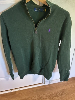 Grön halvzip från Polo Ralph Lauren - Mörkgrön långärmad tröja från Polo Ralph Lauren med halv dragkedja och lila broderad logga på bröstet. Tröjan har ribbade muddar och hög krage. Perfekt för en chill och stilren look.