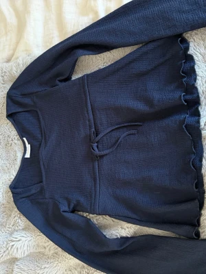 Mörkblå långärmad topp med knyt - Mörkblå långärmad topp med knyt i midjan och volangkant nertill. Toppen har en rund halsringning och är tillverkad i ett mjukt, ribbat material som ger en skön känsla. Perfekt för dig som gillar en enkel men ändå söt stil, nästan helt nytt skick,knappt använd, storlek 146-152.🩵