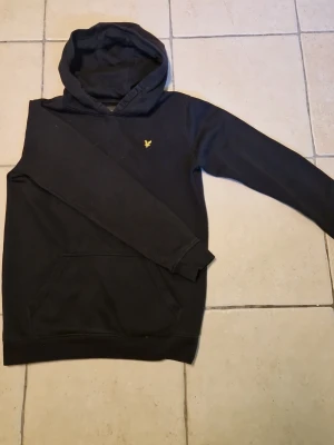 Svart hoodie från Lyle & Scott - Säljer en svart hoodie från Lyle & Scott med klassisk gul logga på bröstet. Hoodien har en stor huva och en praktisk magficka. Materialet är mjukt och bekvämt, perfekt för chill dagar. Långärmad och enkel design som funkar till det mesta.