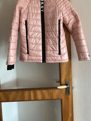 Ljusrosa pufferjacka från DKNY - Snygg ljusrosa pufferjacka från DKNY med svarta detaljer och dragkedjor. Jackan har hög krage, raka ärmar och quiltad design. Perfekt för kalla dagar och har en modern, sportig look med DKNY-logga längs dragkedjan.
