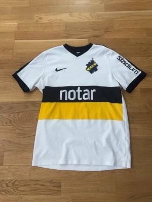 AIK bortatröja Nike med tryck - AIK:s vita bortatröja från Nike med svart och gul rand över bröstet, klubbmärke och sponsortryck. Svart V-ringad krage och svarta detaljer på ärmarna. Nummer 6 och SVEA-tryck på ryggen. Perfekt för fotbollsfans! L MEN ÄR M