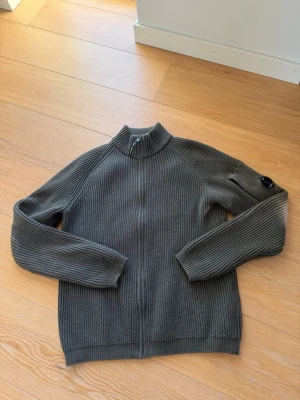 Mörk grön stickad tröja från C.P. Company - Snygg mörk grön stickad tröja från C.P. Company med dragkedja framtill och hög krage. Tröjan har ribbstickad struktur och en ikonisk linsdetalj på ena ärmen. Perfekt för lager-på-lager och streetwear-stil. Stor i storleken