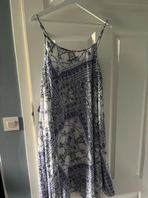 Vit och blå mönstrad sommarklänning - Luftig vit klänning med blått mönster och tunna axelband. Klänningen har en lös passform och är perfekt för varma dagar. Mönstret är inspirerat av boho-stil och tyget känns lätt och svalt mot huden.