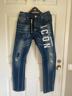 Blå Dsquared2 ICON jeans - Snygga blå jeans från Dsquared2 med stort vitt ICON-tryck och DSQUARED2-logga på ena benet. Jeansen har slitna detaljer och dragkedja fram. Den är perfekt för dig som gillar snygga märkesplagg.