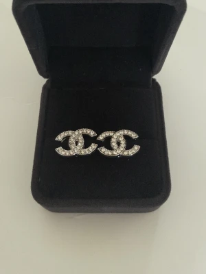 Silver CC örhängen med strass - Snygga örhängen i silverfärgad metall med klassiska CC-loggan täckt av glittrande strass. Perfekt accessoar för att lyfta din stil med en lyxig touch. Levereras i svart ask.