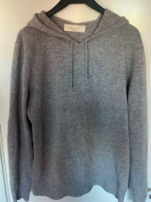 Grå kashmir hoodie från Sayless - Mysig grå hoodie från Sayles i mjuk cashmere. Klassisk modell med huva och dragsnören, samt ribbade muddar vid ärmslut och nederkant. Perfekt för dig som gillar stilrena och bekväma plagg med lyxig känsla.